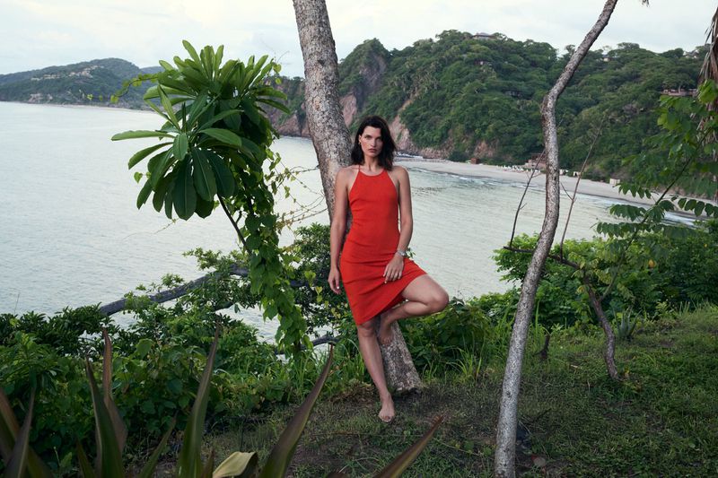 A barefoot woman in a red dress poses against a tree on a tropical coastline, featuring lush greenery, mountains, and ocean views.