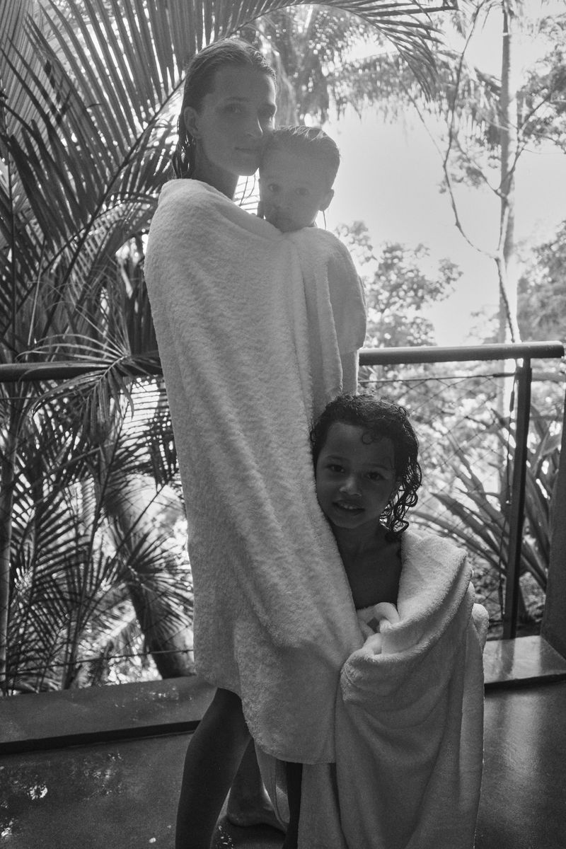 Black and white shot of a mother embracing two children wrapped in white towels outdoors, with palm trees in the background.