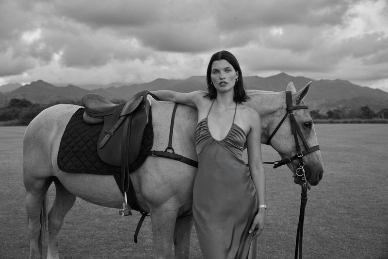 Elegant woman in satin halter dress stands next to horse with saddle in outdoor setting with scenic mountain backdrop, black and white.