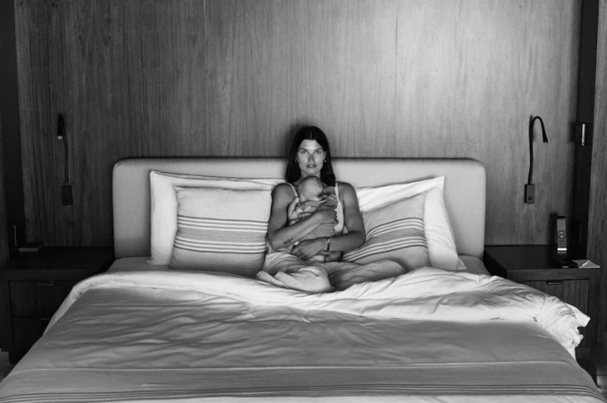 Black and white image of woman seated on a bed, cradling a baby, with natural light highlighting the serene indoor scene.