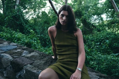 Model in an olive green dress sitting on a low stone wall amidst a lush tropical forest environment.