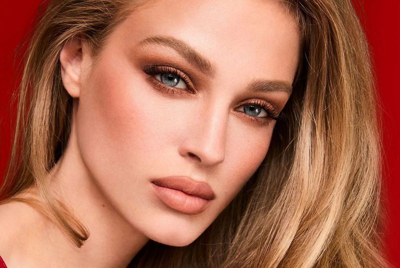 Close-up portrait of a young fair-skinned woman with blonde hair, wearing brown eyeshadow and nude lipstick, against a red background.