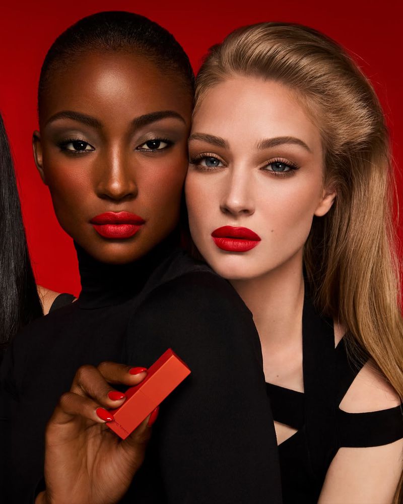 Two women pose for a beauty shot with bright red lipstick, holding product against a red backdrop. Fashion and cosmetic advertising.