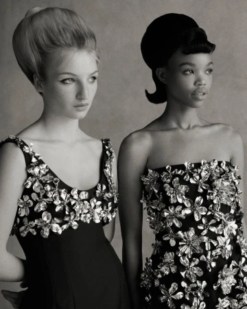 Two women with stylized retro hairstyles and dresses with floral embellishments, captured in a monochrome portrait, fashion-forward.