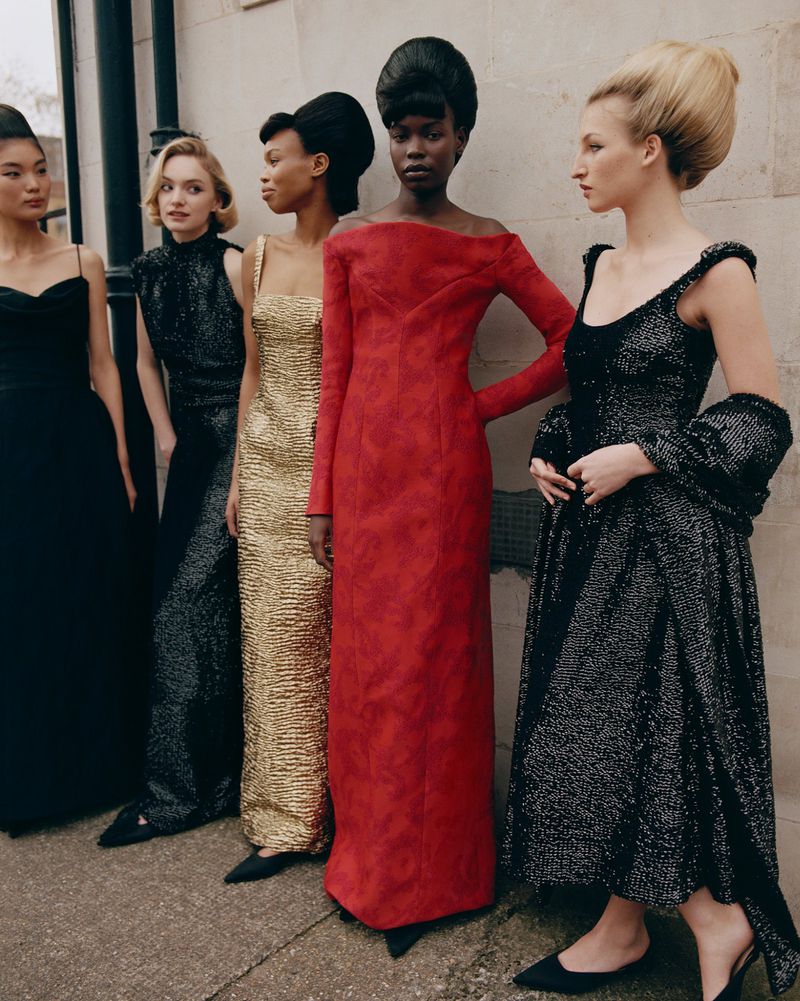 A group of five women standing together wearing formal gowns with vintage hairstyles in front of a beige, textured wall.