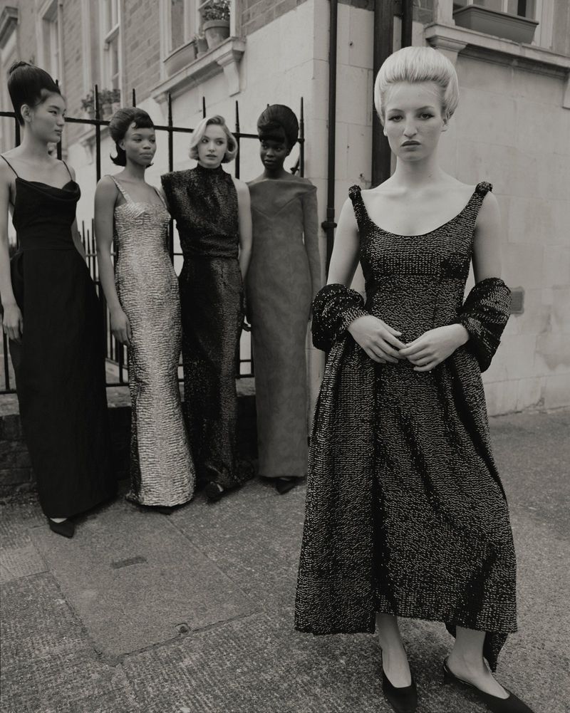 Black-and-white fashion shot features five women in formal gowns outdoors. The model in the front is in focus, while others are blurred.
