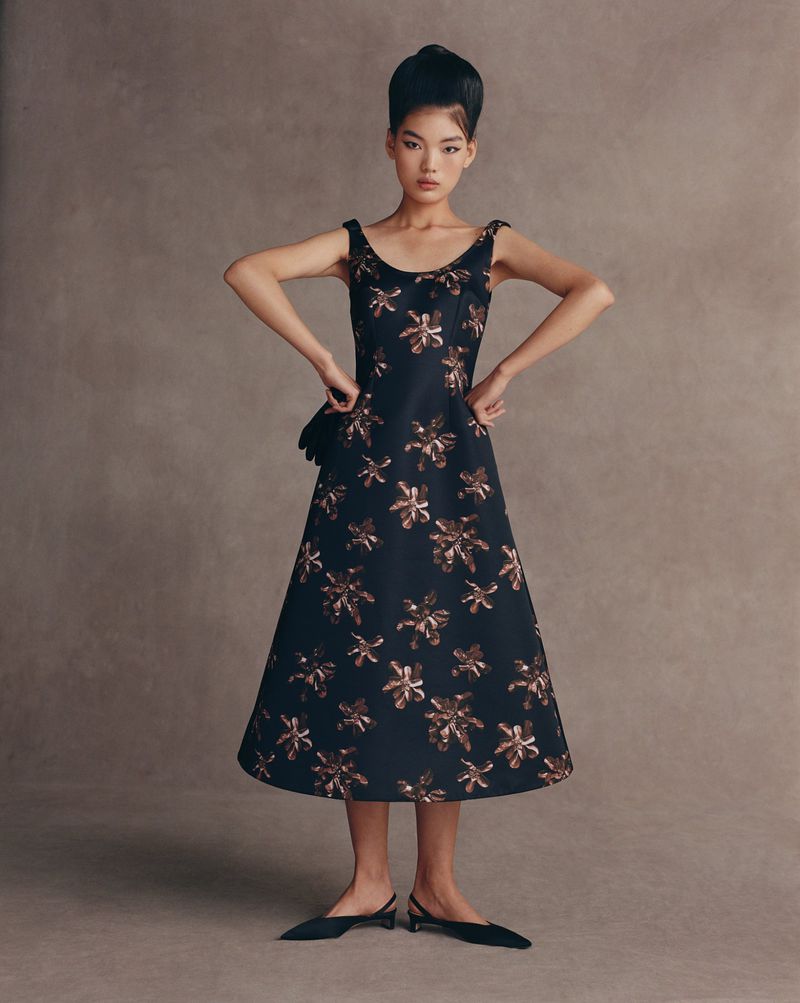 Young woman poses in a vintage style sleeveless black A-line dress with gold floral pattern and black slingback heels against a neutral backdrop.