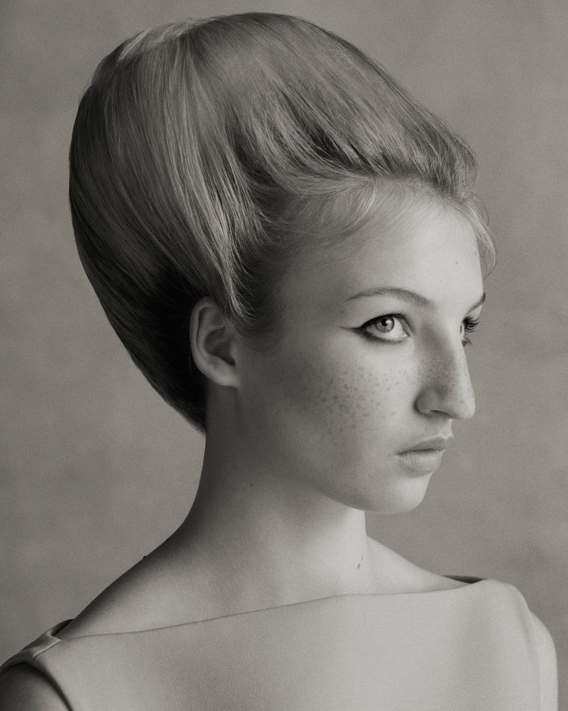 Monochrome portrait of a young woman with freckles, wearing a simple garment, and featuring a retro hairstyle and distinctive eyeliner.