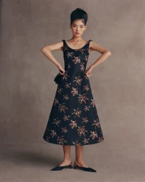 East Asian model in a black metallic floral A-line midi dress and black slingback flats against a warm textured studio backdrop.