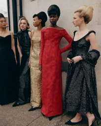 Five fashion models in evening gowns including red, gold, and black dresses posing together against a stone exterior wall.