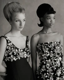 Two models with retro 1960s bouffant hairstyles wearing black dresses adorned with metallic floral embellishments against a studio backdrop.