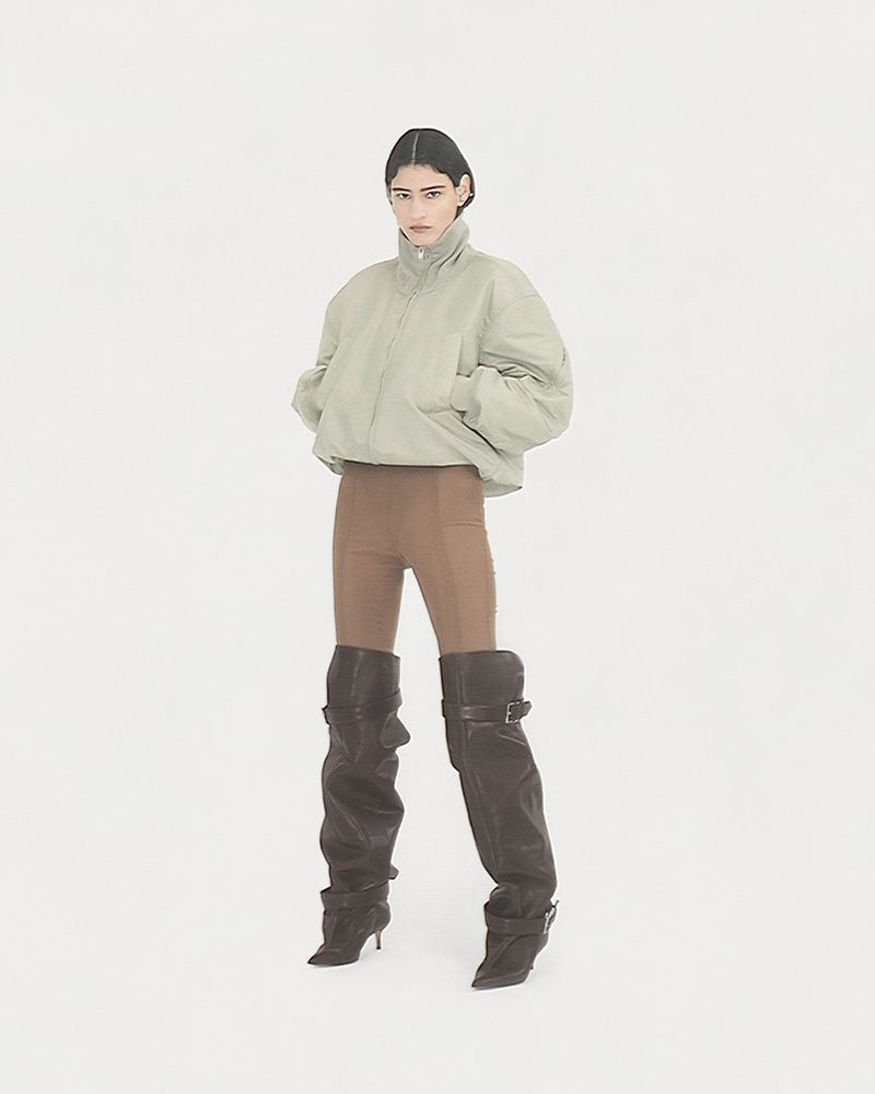 A model poses in a studio wearing a light green puffer jacket, brown leggings, and black leather heeled boots against a white background.