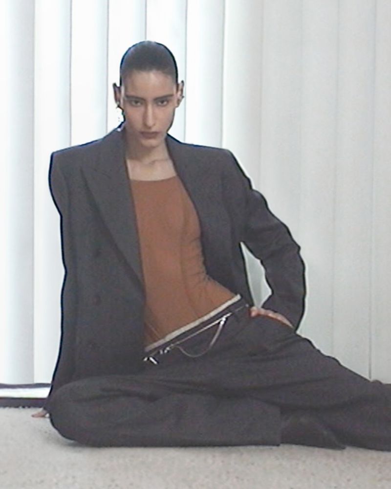 A woman in a gray suit poses on the floor in front of blinds in a fashion portrait, looking directly at the camera with neutral expression.