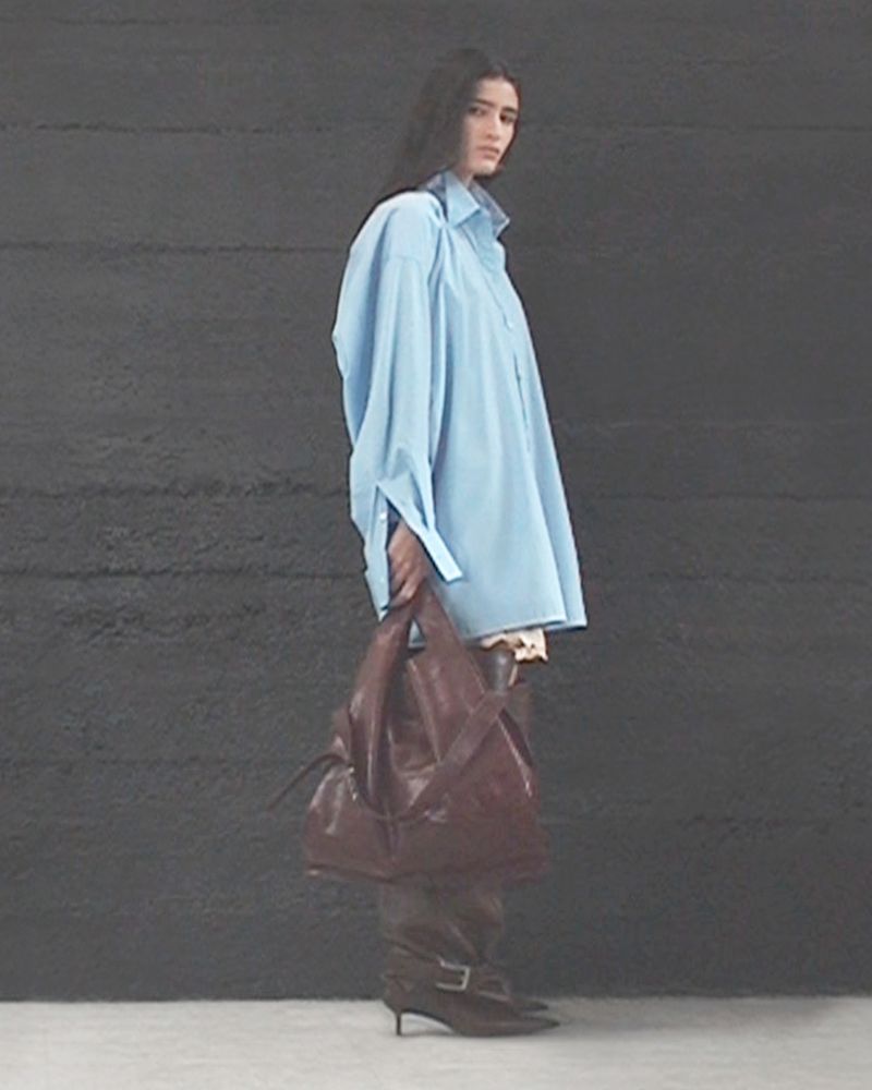 Fashionable adult female posing in blue oversized shirt, high-heeled boots, and holding a brown leather handbag against a dark backdrop.