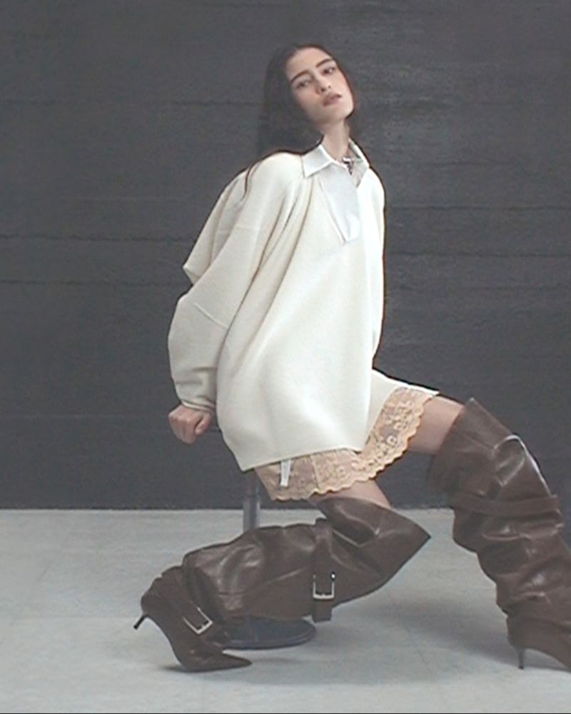 A brunette woman sits on a stool in a studio, wearing an oversized cream sweater, lace trim slip, and brown high-heeled boots.