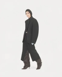 Model Luiza Perote poses in an oversized black jacket, black skirt, and slouchy dark leather heeled boots against a white backdrop.