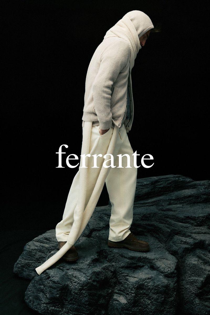 Man in textured hooded sweater and white pants poses on a stone formation against a dark backdrop, fashion editorial, Ferrante branding.