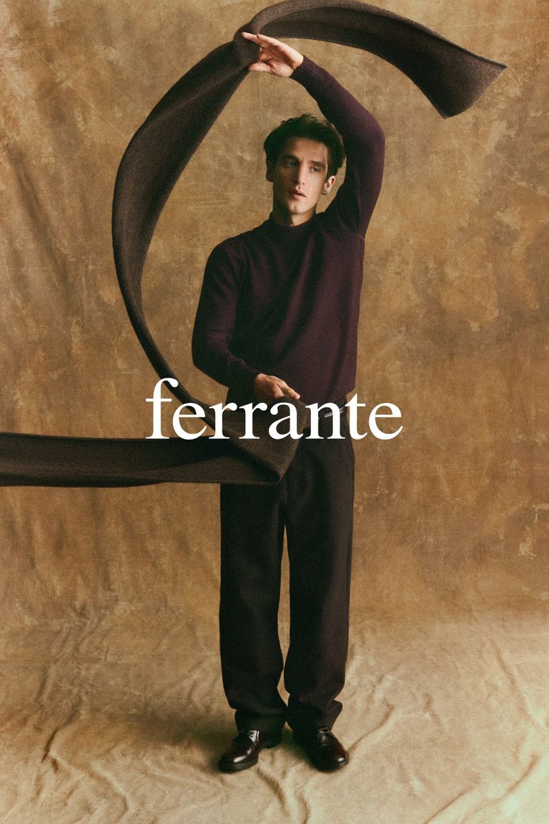 A slender man in a purple sweater and dark pants poses with a draped brown scarf against a textured brown background in a studio shot.