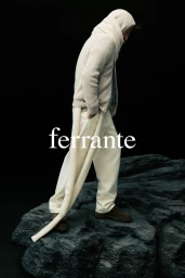 Model Anatol Modzelewski standing in cream knitwear and trousers on dark textured rocks against a black backdrop.