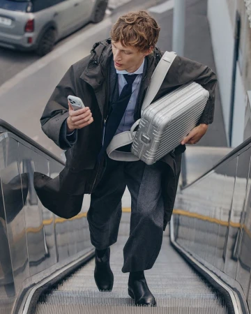 Man in a trench coat and carrying a silver briefcase on an escalator while looking at his smart phone in an urban setting.