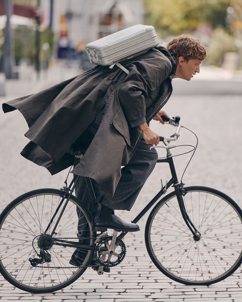 An adult male rides a black bicycle through an urban setting. He wears a long coat with a silver suitcase strapped to his back.