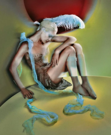 Artistic studio shot of a fair-skinned adult woman in a metallic dress with blue ribbons and a feather headpiece sitting in a corner.