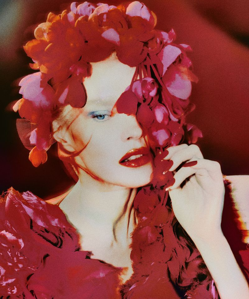 Close-up portrait of a person with light skin and blue eyes, adorned with red floral headwear and makeup, set against a red background.