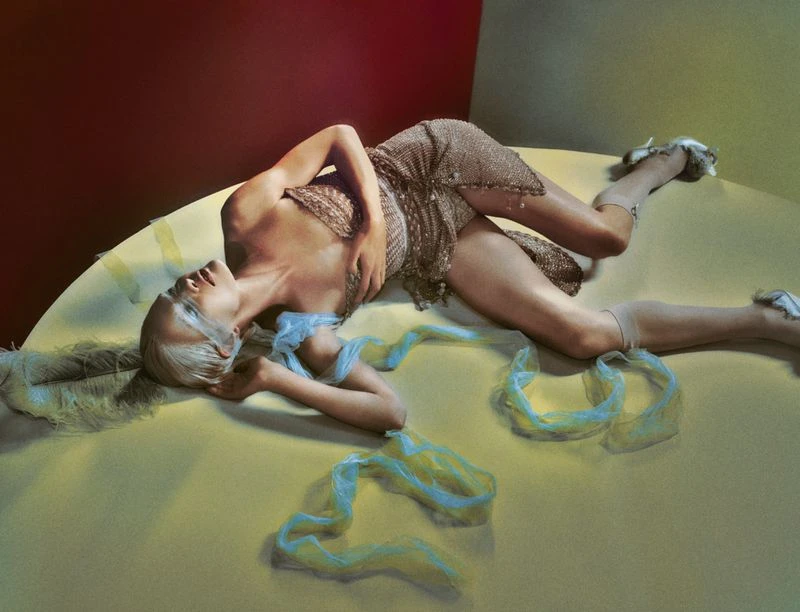 A woman with blonde hair is lying down in a dress, with blue ribbon accents, in a studio setting with red and tan background.