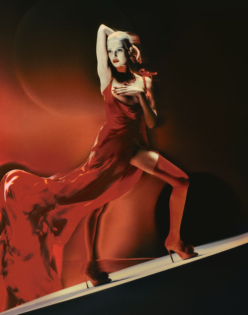 Elegant woman in red dress and heels poses on a white surface with red, orange, and black background in a fashion photography style.