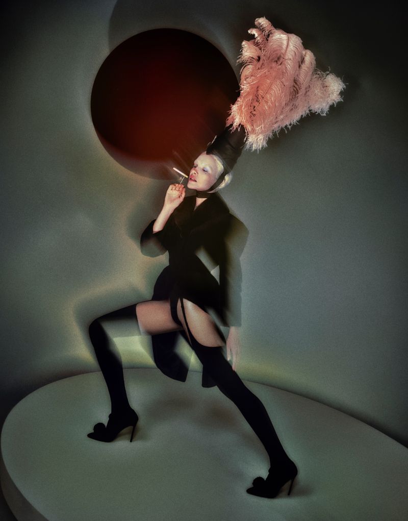 Fashionable adult female model poses in studio wearing a dark jacket, thigh-high stockings, heels, and elaborate feathered hat while smoking.