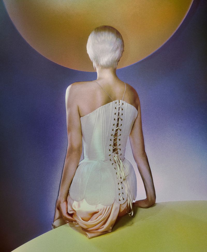 Back view of a woman with short blonde hair wearing a white corset dress sitting on a yellow surface in front of a gradient background.