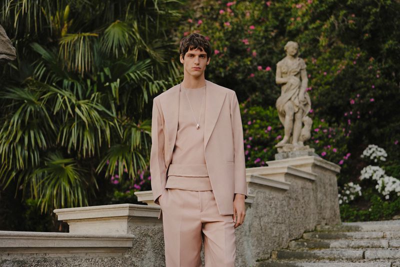 A stylish adult male poses outdoors on stone steps in a blush suit and matching t-shirt, accessorized with a pendant necklace.