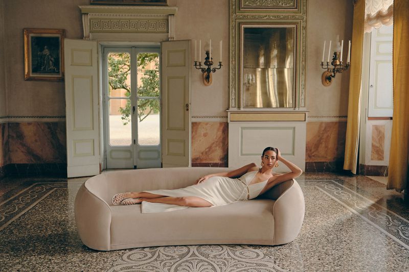 An adult female lounges on a sofa in a light dress indoors, accessorized with jewelry, set against an ornate backdrop of doors and walls.