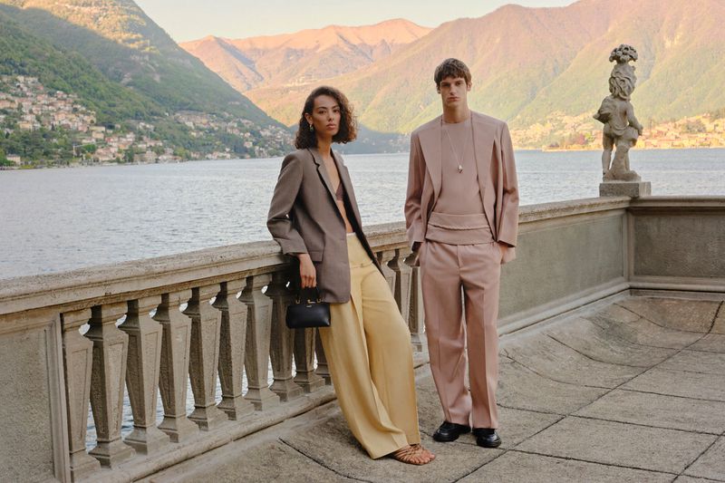 Two fashion models pose outdoors in formal wear overlooking a scenic lake and mountain landscape backdrop on a sunny day.