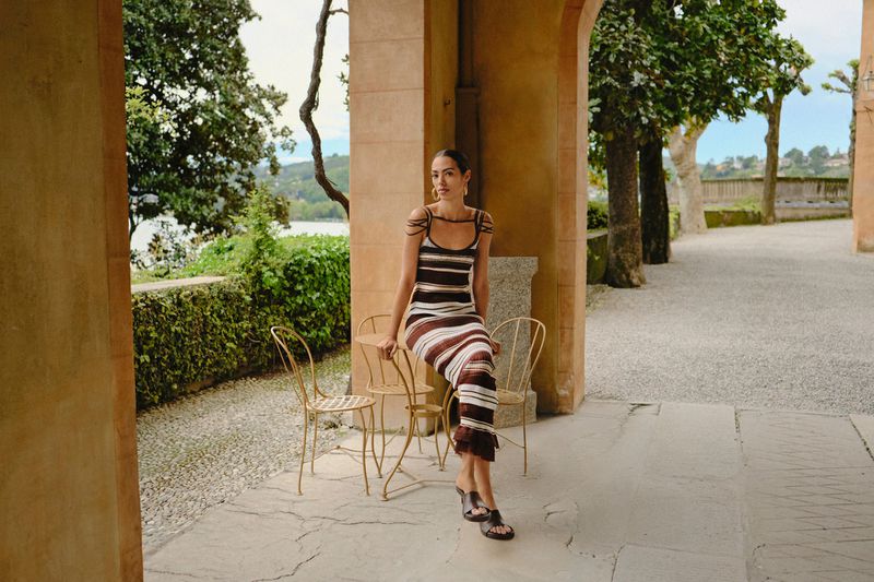 A woman poses sitting on a gold metal chair outdoors under a stone archway, wearing a striped knit dress and black slide sandals.