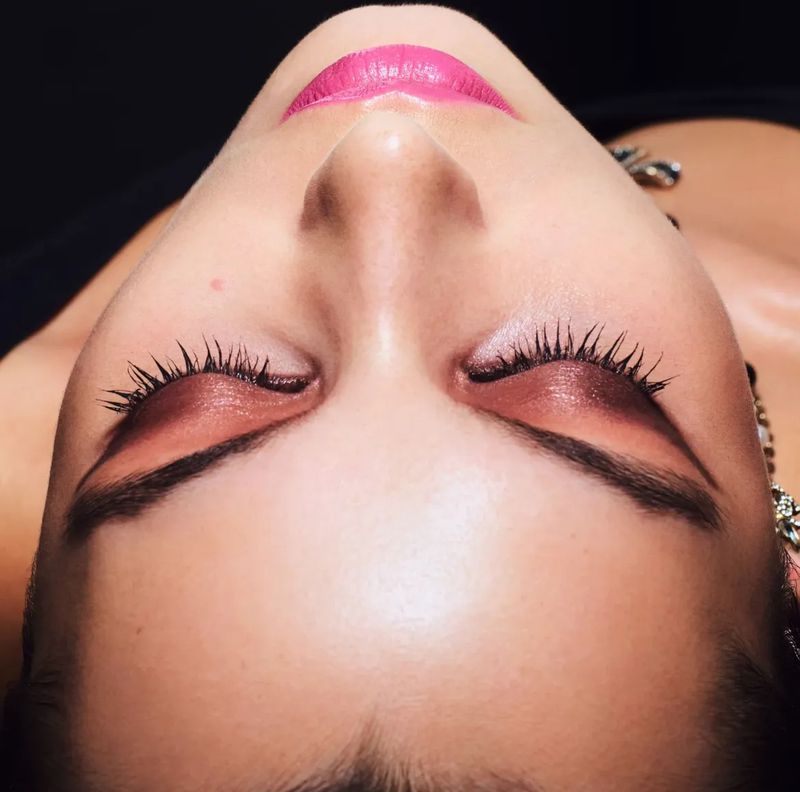 Close-up of a woman's face with pink lipstick and brown eyeshadow. The shot is from directly above her face. Dark background.