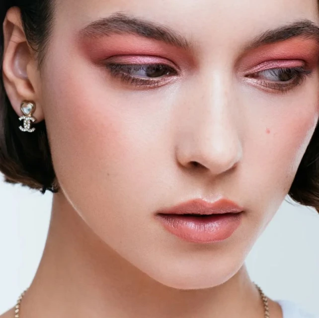 Close-up portrait of a woman with red eyeshadow, glossy lips, and Chanel earrings against a light grey background, showcasing beauty and fashion.