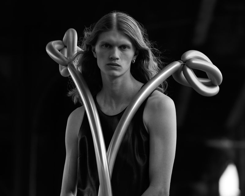 Monochrome portrait of a slender adult with long blond hair wearing a tank top and holding balloon art in front of a dark background.