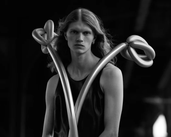 Monochrome portrait of a slender adult with long blond hair wearing a tank top and holding balloon art in front of a dark background.