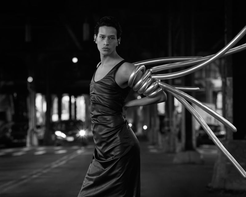 Person in black dress with surreal silver arm extensions poses on urban street at night in dramatic black and white fashion photo.