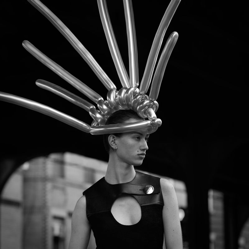 Person wearing a black outfit and a silver balloon headpiece stands against an urban backdrop in this black and white fashion shot.