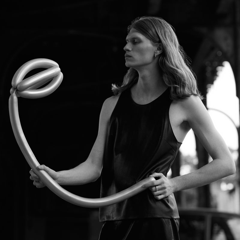 Monochrome portrait of a slender adult with long hair holding a twisted balloon against a blurred urban background.