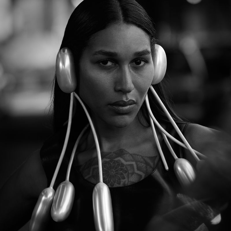 Black and white portrait of an adult female with long dark hair wearing surreal balloon earrings in a blurred indoor environment.