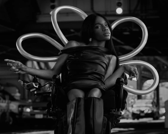 Black and white portrait of a confident woman in a wheelchair, wearing a dress and boots, posed against an urban background.