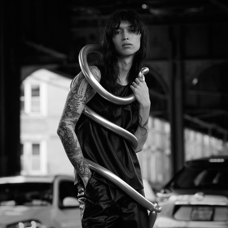 Monochrome portrait of a tattooed person with long hair wearing a satin top and silver balloon necklace, under an overpass with vehicles.