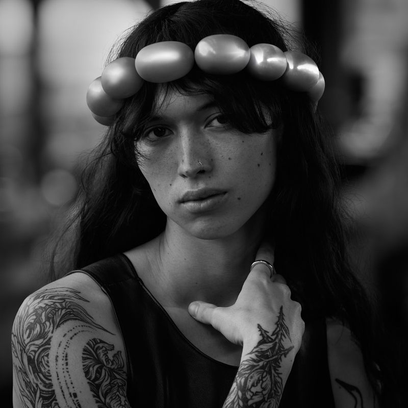 Portrait of a woman with dark hair and tattoos wearing a beaded headpiece, against a blurred indoor background in black and white.