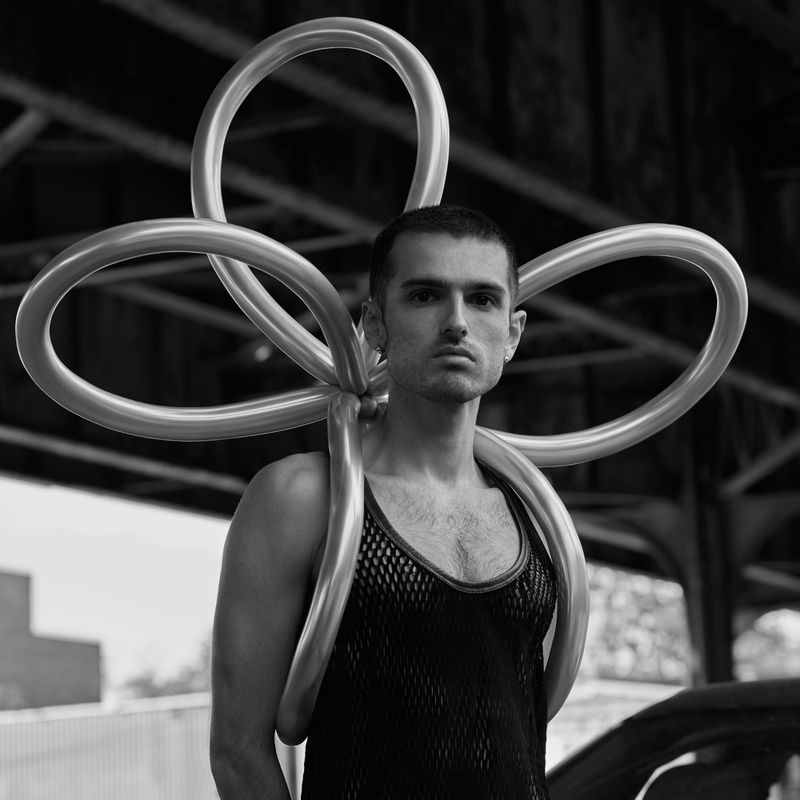 Monochrome portrait of a man wearing a mesh top and a balloon structure over his shoulders, standing outdoors under a structure.
