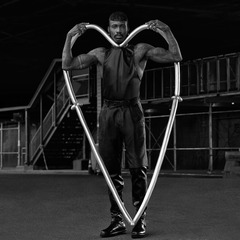 Adult male posing with a heart-shaped metal frame wearing a sleeveless top, pants, and boots in an industrial location in black and white.