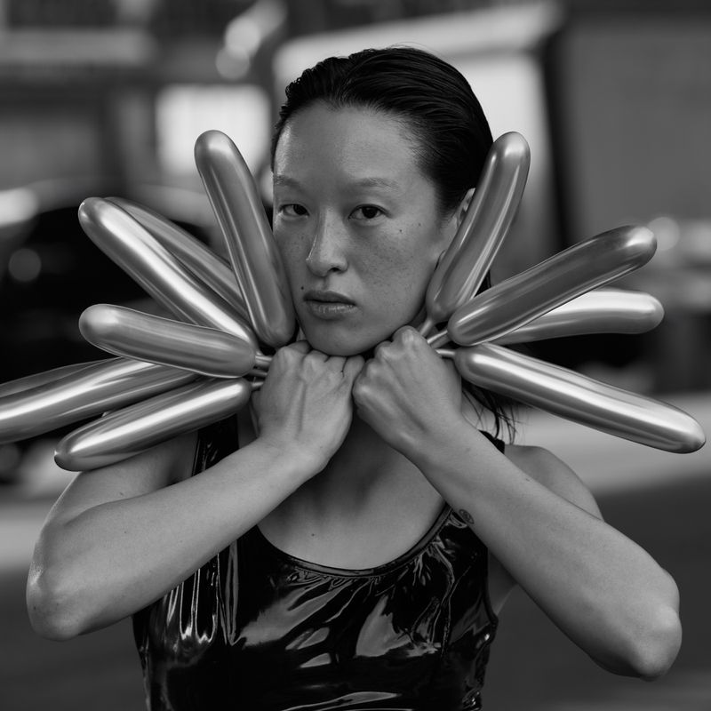 Monochrome portrait of a young Asian woman with dark hair slicked back, shiny black top and metallic balloon collar in an urban setting.