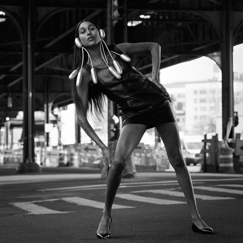 Black and white fashion shot of a woman in a dress and heels posing with unique futuristic jewelry under a bridge in a city.